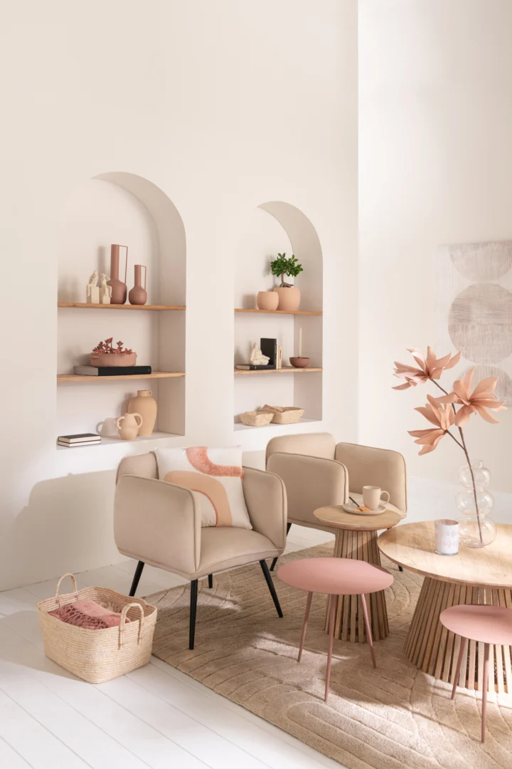 Een moderne, neutraal getinte woonkamer met beige fauteuils, Tafel Vincent Mangohout Natuurlijk Medium als de ronde houten tafel, decoratief roze keramiek op gewelfde wandplanken, een geweven mand, blush accenten en gedroogde bladeren in een vaas voor een natuurlijke touch.