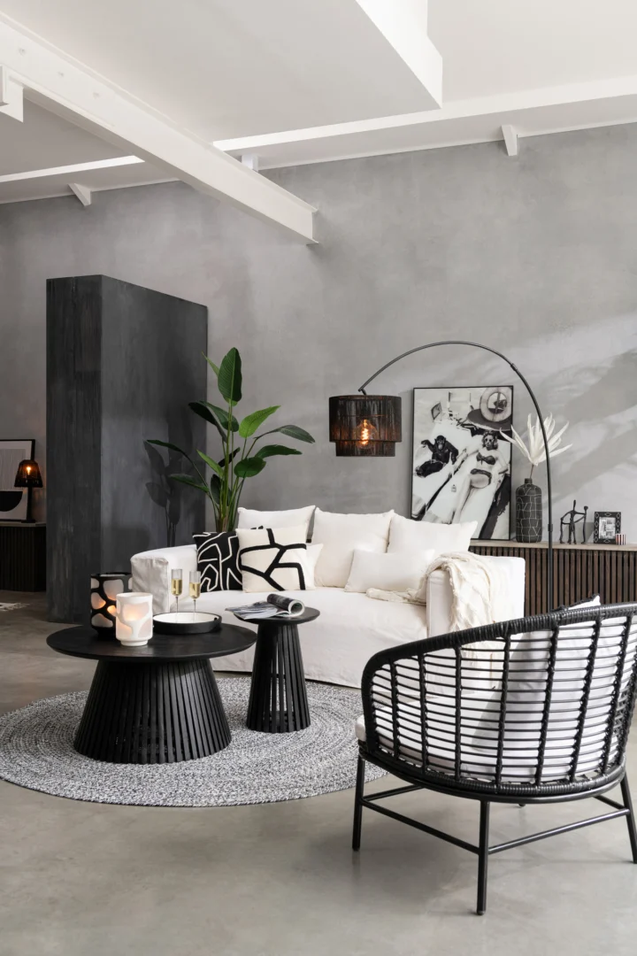 Een moderne woonkamer met een witte bank, monochrome kussens, een rond grijs vloerkleed, twee Tafel Vincent Mangohout Zwart Middelgroot salontafels, een geweven stoel, een potplant en decor tegen een grijze muur.