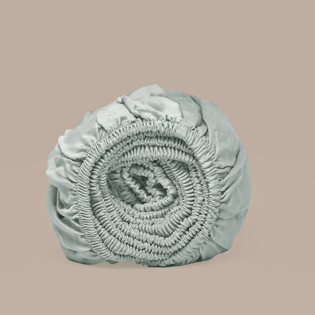 Oprolbaar groen topper hoeslaken van Tencel satijn. Zachte stof met elastiek, gerold op een beige achtergrond. Zachte belichting.