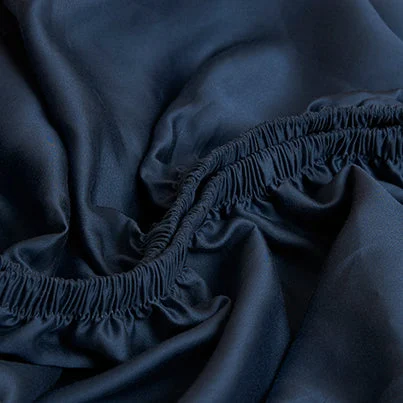 Close-up van een blauw TENCEL hoeslaken met elastiek. De stof heeft een subtiele glans en zachte plooien. Zachte belichting accentueert de textuur.