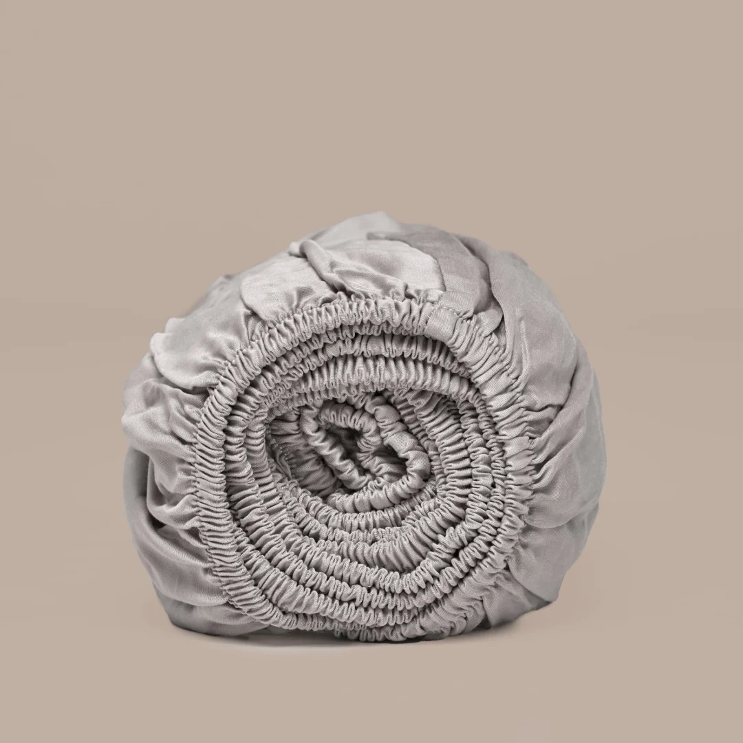 Oprolbaar taupe hoeslaken van Tencel™ satijn. Zachte stof met elastiek, strak opgerold tegen een beige achtergrond.