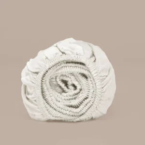 Opgerold, off-white TENCEL™ satijnen hoeslaken, close-up. Zachte belichting, effen beige achtergrond. Textuurrijk, luxe uitstraling.