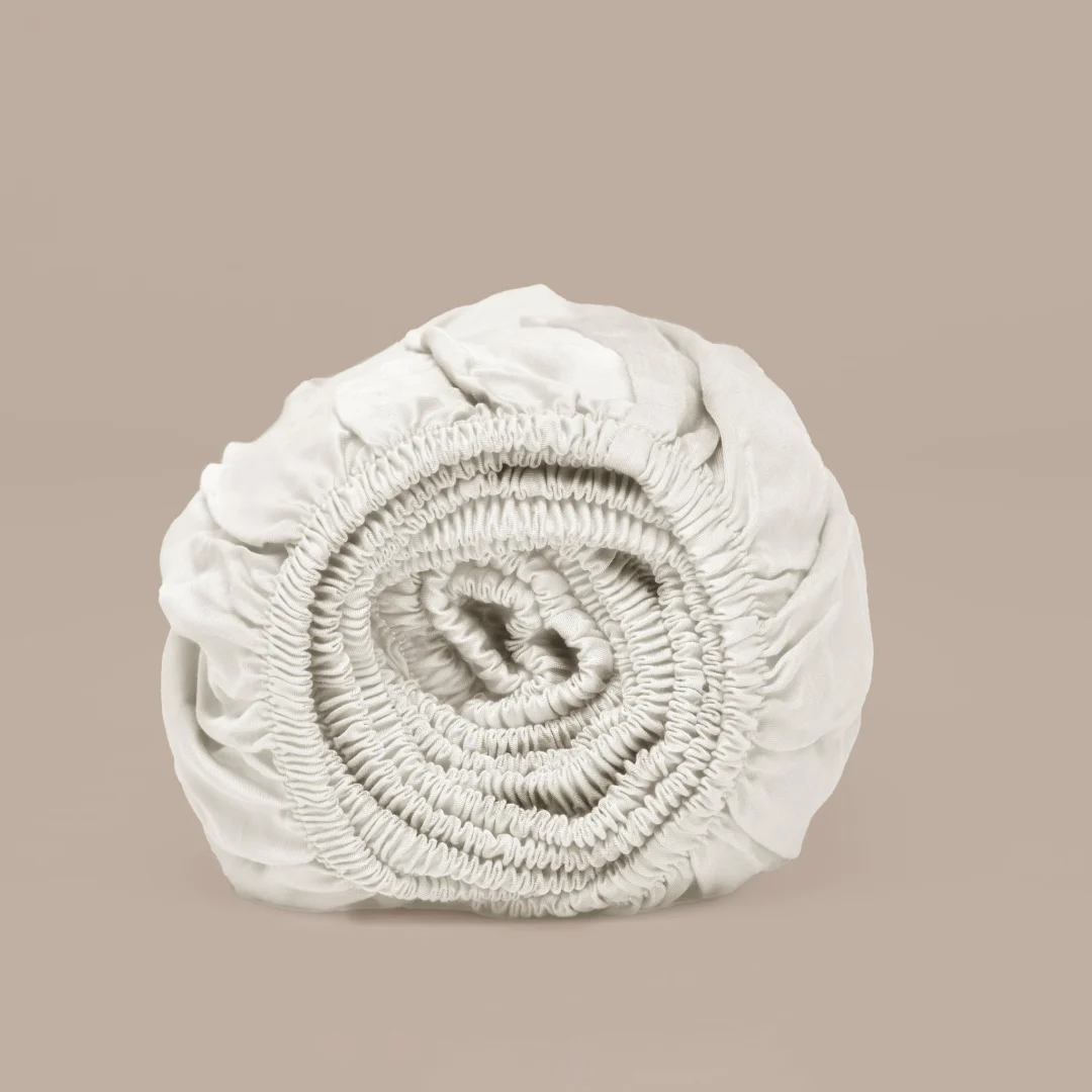 Opgerold off-white hoeslaken van Tencel™; zachte stof, elastische randen. Neutrale achtergrond, zacht licht. Detailopname van textuur en materiaal.