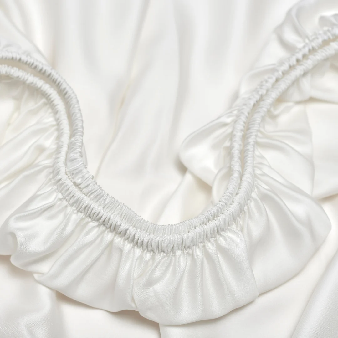 Close-up van een off white hoeslaken met elastiek, zachte stof. Lichte schaduwen geven diepte aan het gladde, ademende Tencel™ materiaal.