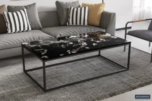 In een moderne woonkamer met een grijze bank en gestreepte kussens staat de Salontafel Zwart Marmer Minimalistisch, een rechthoekige zwart marmeren salontafel met een strak metalen frame, op een grijs vloerkleed met textuur.