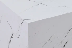 De Bijzettafel Wit MDF Design heeft een geometrische blokvorm met een wit marmerachtig oppervlak, geaccentueerd door fijne natuurlijke grijze aderen en schuine randen, tegen een bijpassende wit marmeren achtergrond.