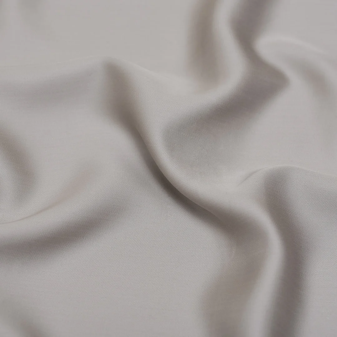 Close-up van een taupe hoeslaken in TENCEL™ satijn. De stof is zacht golvend en het licht valt er subtiel op. Ademend materiaal in neutrale tint.