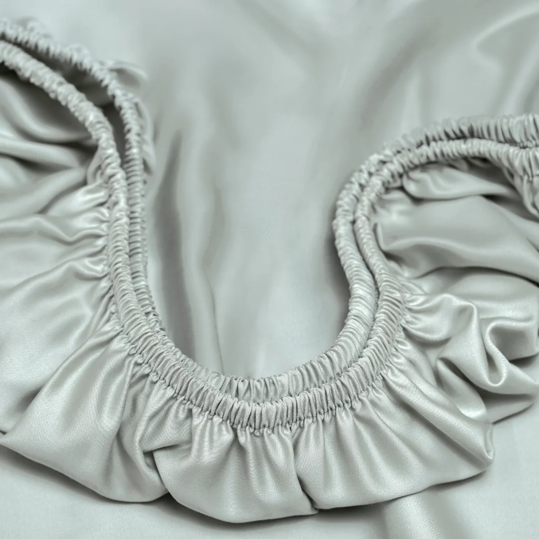 Close-up van een grijsgroen TENCEL™ hoeslaken met elastiek. Zachte stof met subtiele glans en plooien, vanuit een hoog camerastandpunt.