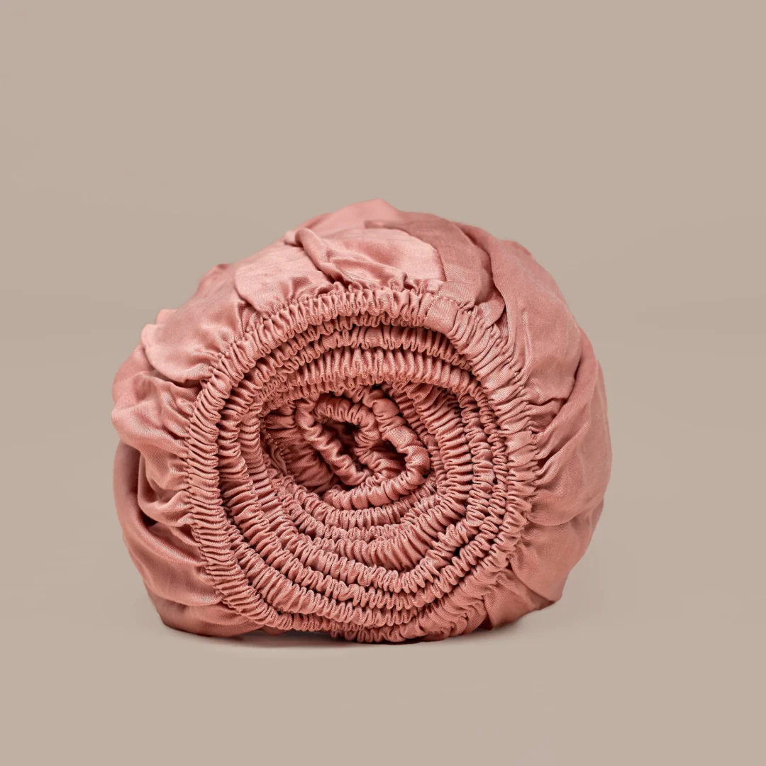 Oprol van een terra roze hoeslaken van TENCEL™ Lyocell satijn. Zachte plooien en elastiek zichtbaar. Neutrale achtergrond, zachte belichting.