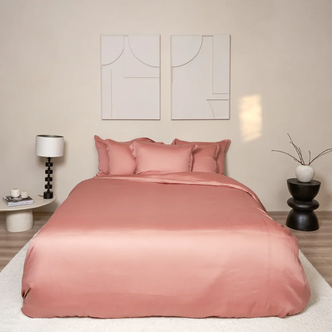 Terra roze satijnen beddengoed opgemaakt op een bed met een wit vloerkleed. Zachte belichting accentueert de luxe uitstraling van het hoeslaken.