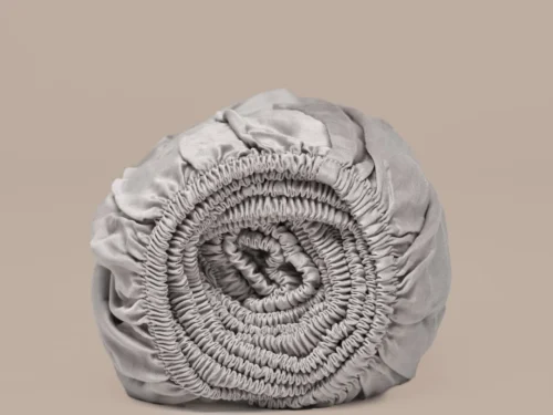 Oprolbaar taupe hoeslaken van satijn TENCEL™, zijdezacht. Zachte, neutrale achtergrond. Elastische randen zichtbaar in detail.
