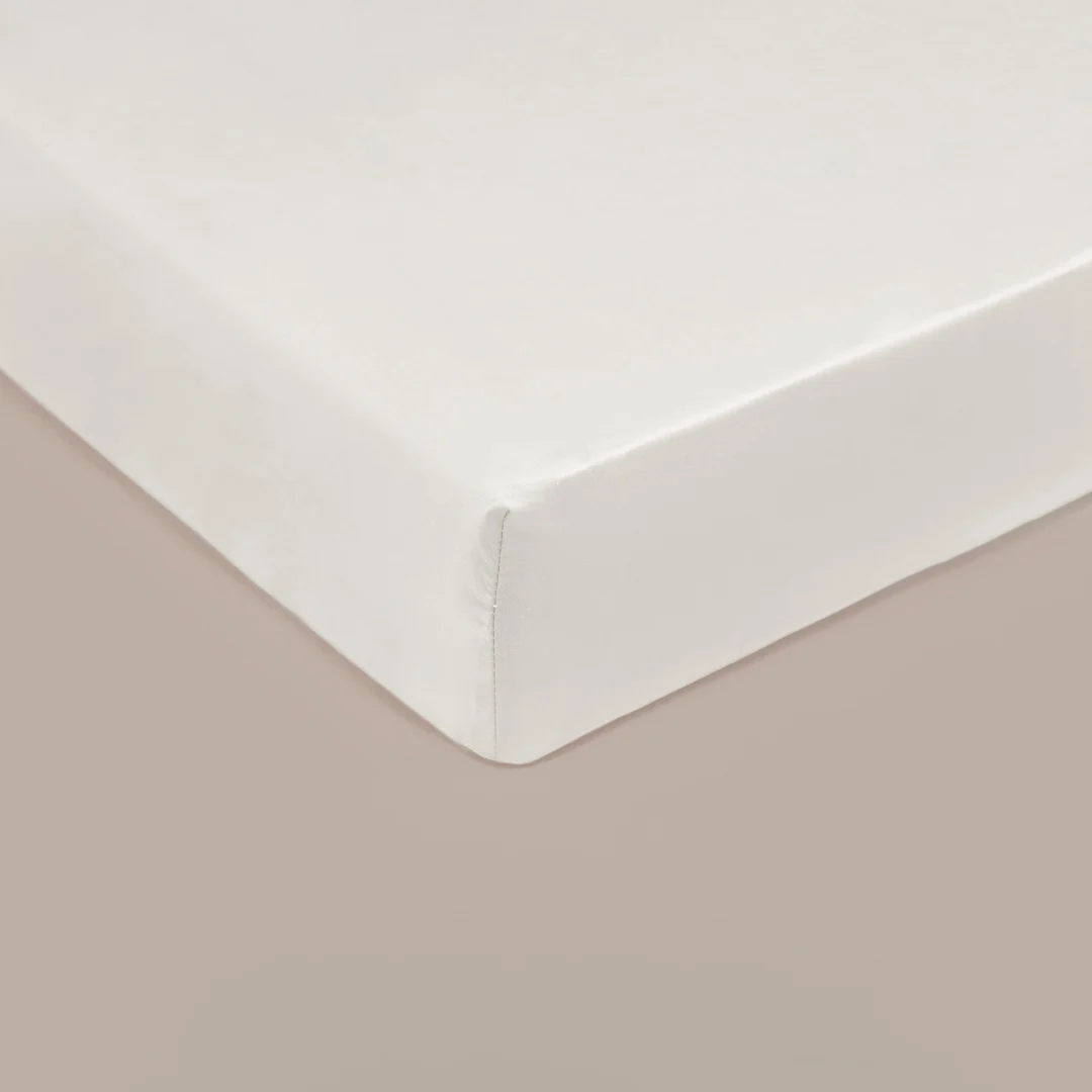 Detailopname van een off-white hoeslaken. Satijnzachte stof, strakgetrokken over een matras. Zachte belichting, warme beige achtergrond.