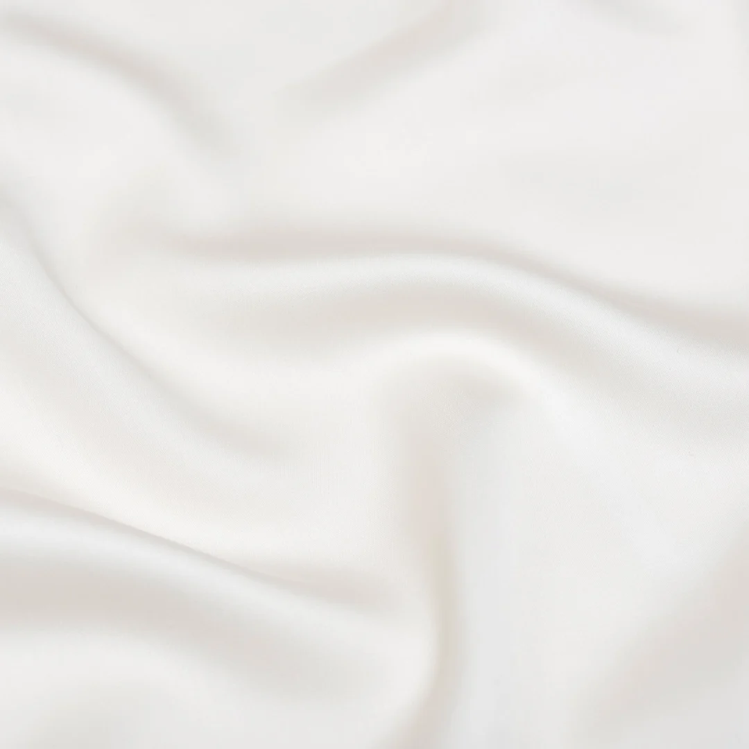 Close-up van een off-white hoeslaken met zachte satijnglans. De stof golft subtiel onder zacht, diffuus licht. Textuurrijk en uitnodigend.