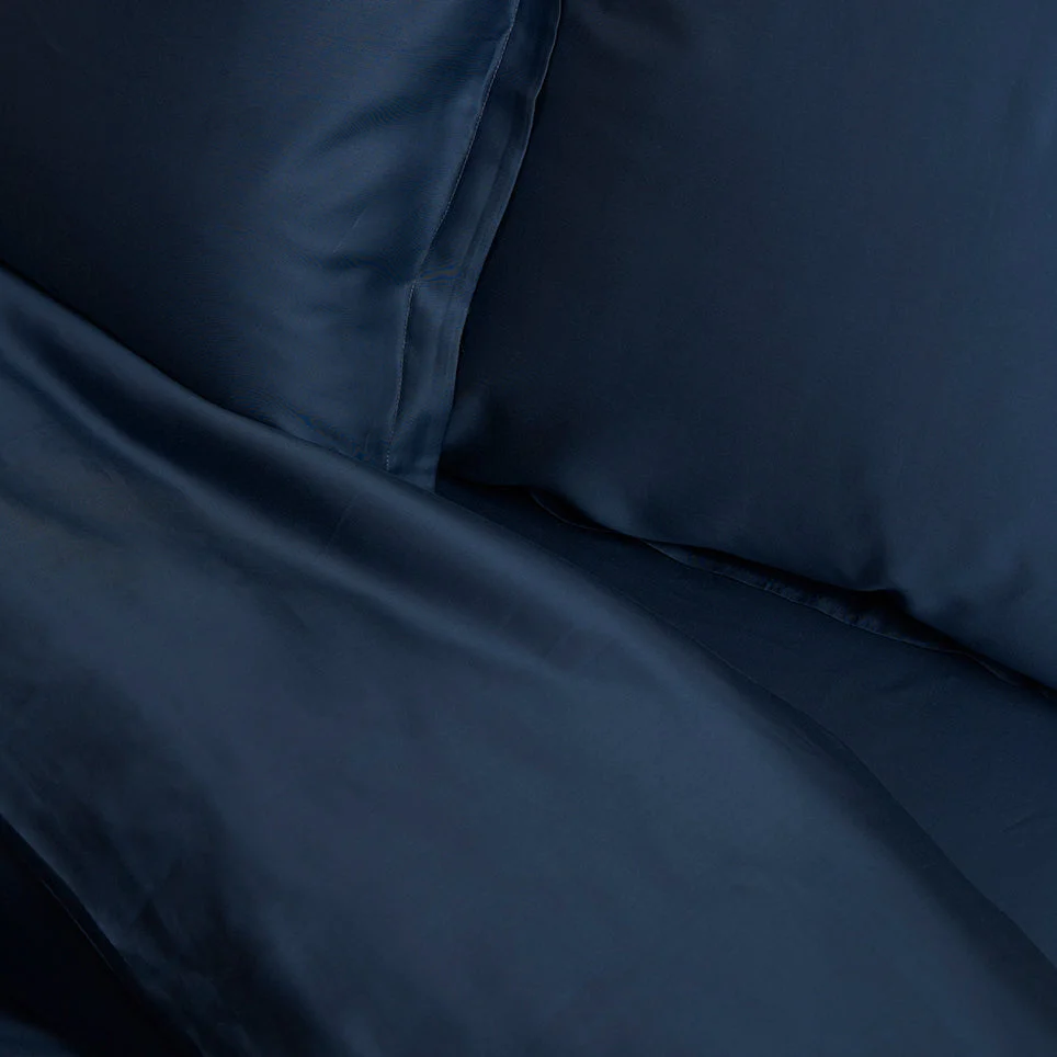 Close-up van een blauw hoeslaken. De stof is glad en glanzend, met zachte plooien. Het licht valt subtiel op het TENCEL lyocell materiaal.