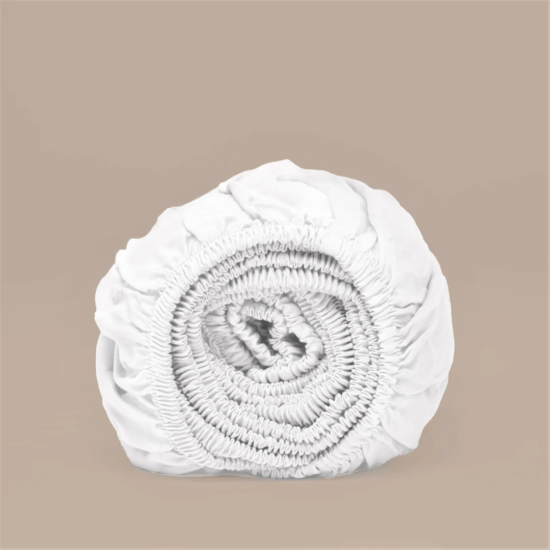 Rol van wit hoeslaken van TENCEL™ Lyocell satijn, zachte stof. Neutrale achtergrond, zachte belichting. Elastische randen zichtbaar.