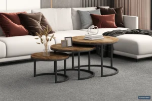 Een moderne woonkamer heeft een Salontafel Roest Mangohout Design, een trio ronde houten salontafels met zwarte metalen poten op een grijs tapijt, versierd met een glazen sculptuur, ingelijste foto's en een vaas met gedroogde planten naast een lichtgrijze sofa.