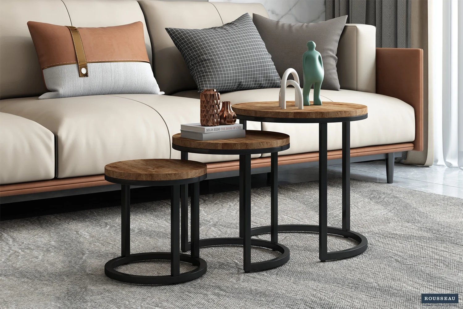 De Bijzettafel 'Plano' 3-delig Mango hout set - drie ronde houten nesttafels met zwart metalen frames - staat voor een beige en bruine bank met gedessineerde kussens, bekleed met decor en op een grijs gedessineerd vloerkleed.