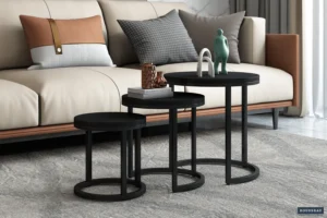 Drie moderne zwarte 'Plano' 3-delige mangohouten bijzettafels staan voor een beige bank met grijze en bruine kussens. Op de tafels staan decoratieve voorwerpen en eronder ligt een tapijt met een grijs patroon.