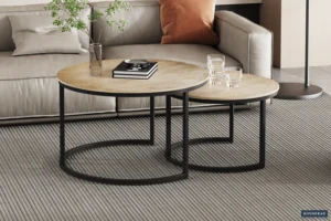 De Salontafel Zilver Compact, met twee ronde tafels met een beige blad en een zwart metalen onderstel, staat op een grijs gestreept vloerkleed voor een lichtgrijze bank. De tafels zijn gestyled met een glazen vaas, boek en drie glazen.