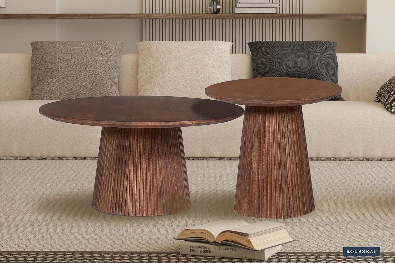 De Salontafel 80 CM Rond Mangohout met een geribbeld onderstel staat op een licht getextureerd vloerkleed voor een beige bank met zwarte kussens en kussens met patronen. Een open boek rust op het tapijt op de voorgrond.