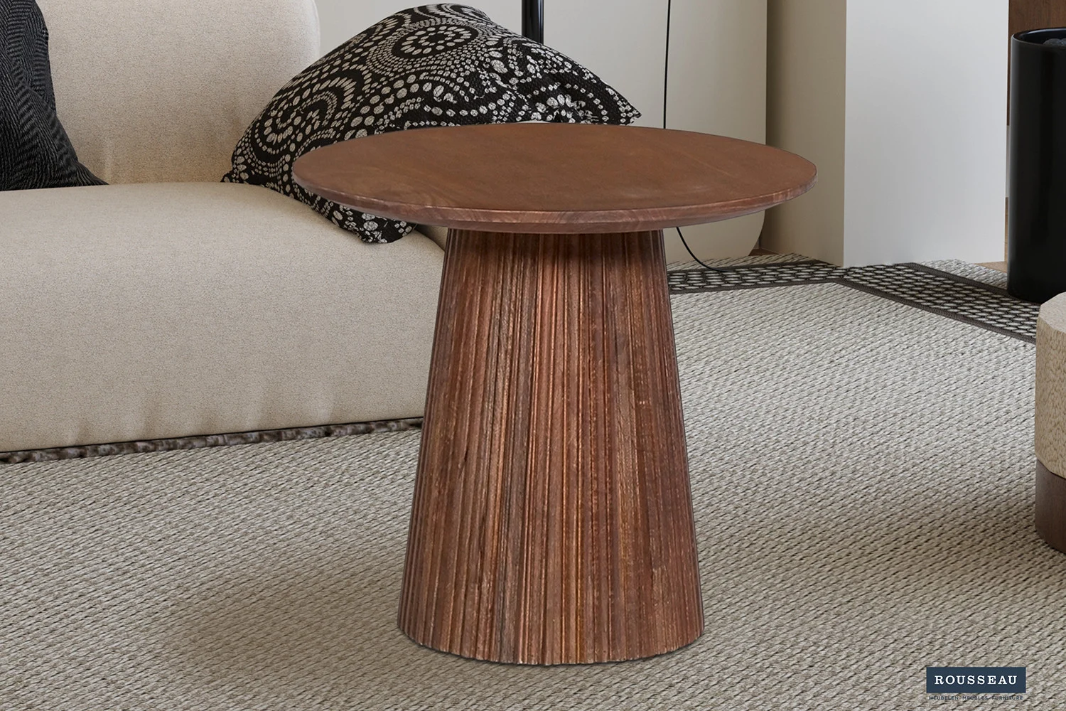 De Salontafel 50 CM Rond Mangohout Design met een geribbelde basis rust op een licht getextureerd tapijt naast een beige bank met daarop een kussen met zwart-wit patroon.