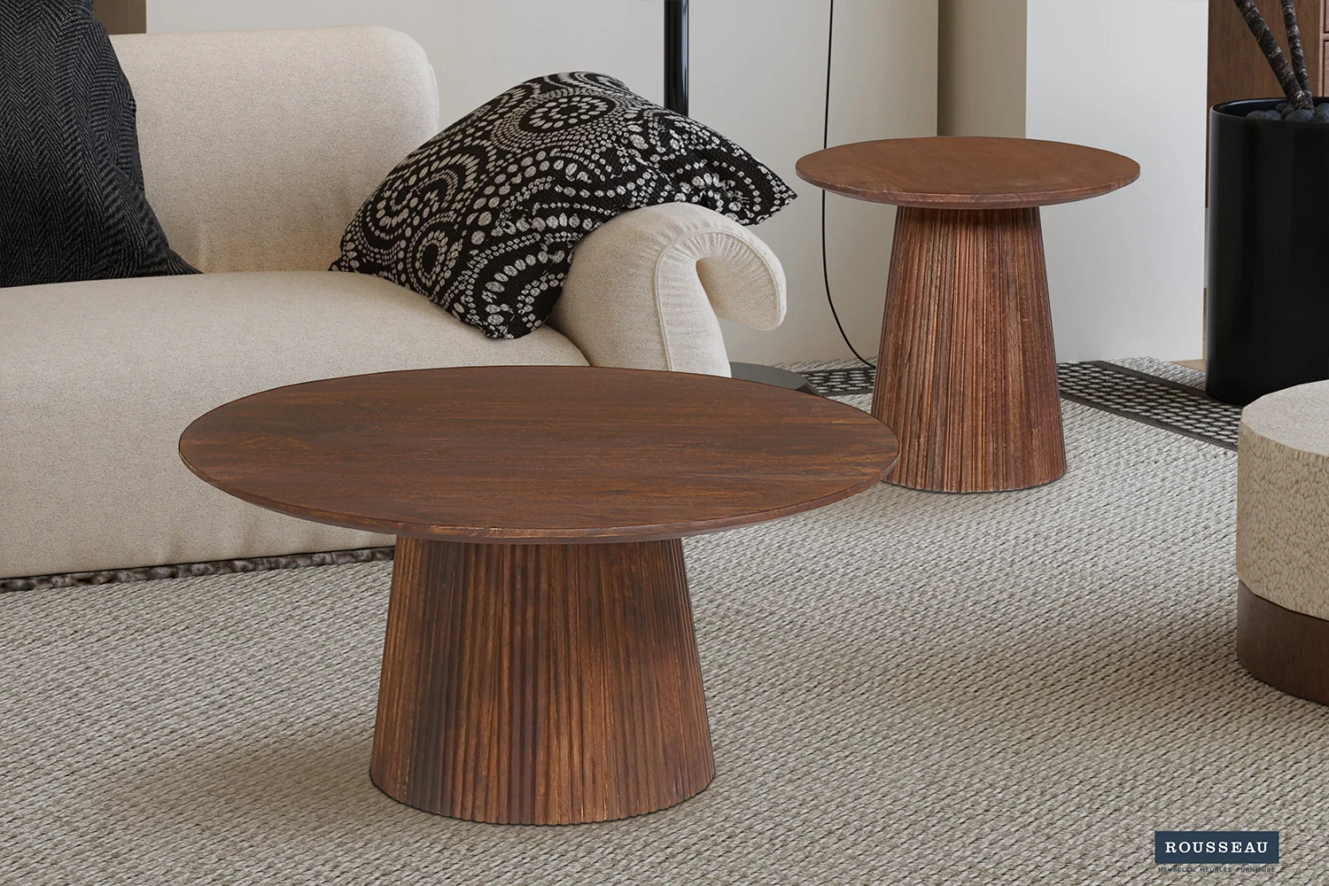 Een moderne woonkamer met een beige sofa, zwart-witte kussens met patroon en twee Salontafel 50 CM Rond Mangohout Design salontafels met cilindrische onderstellen en platte bladen op een beige vloerkleed met textuur.