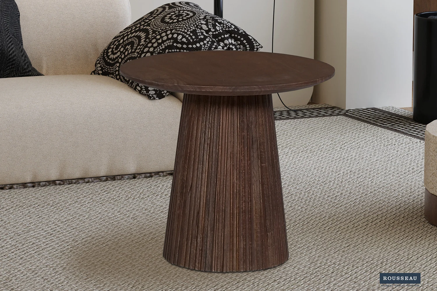 De Salontafel 50 CM Rond Mangohout Modern, een ronde bijzettafel van donker hout met een geribbelde conusvoet, rust op een licht getextureerd tapijt naast een beige bank en een kussen met zwart-wit patroon.