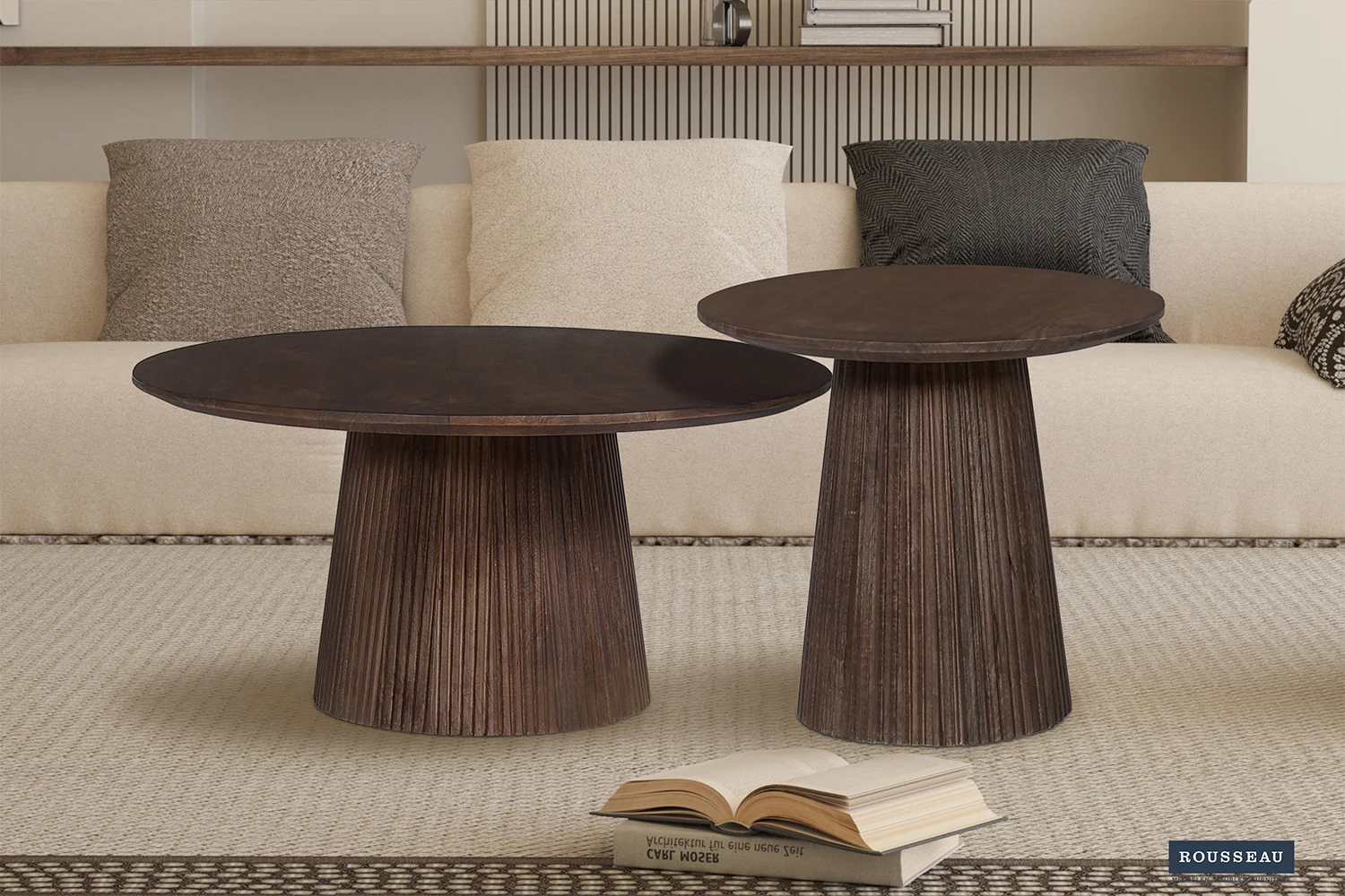 Een Salontafel 50 CM Rond Mangohout Modern met een geribbeld onderstel staat op een vloerkleed met een licht patroon voor een beige bank met neutrale en donkere kussens. Een open boek ligt op de grond naast de tafel.