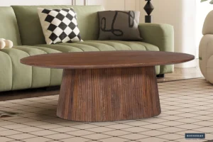 De Salontafel 120 CM Ovaal Mangohout, een klassieke ovalen salontafel van mangohout, staat op een gedessineerd vloerkleed voor een groene bank met zwart-witte geometrische kussens in een moderne woonkamer.