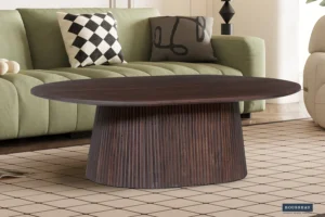 Een moderne woonkamer met een groene bank, twee gedessineerde kussens en de Salontafel 120 CM Ovaal Mangohout Design als een donkerhouten salontafel op een beige vloerkleed met rasterpatroon. Een lamp en abstracte kunst verschijnen op de achtergrond.