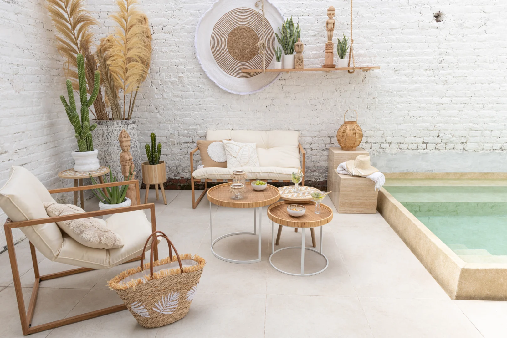 Een lichte, moderne patio met een klein zwembad, houten meubels, planten in potten, boho-chique accenten en een Set van 3 bijzettafel patronen 3 poten Bamboe/Hout Natuur/Wit tegen een witte bakstenen muur.