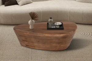 De Salontafel Mangohout Industrieel Modern, een ovale houten salontafel, staat op een beige vloerkleed met een dienblad met een fles, glas en schaal naast een vaas met gedroogd blad, voor een lichtgekleurde sofa.