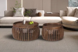 Twee Salontafel Roest Mangohout Design ronde tafels met open lattenbodems staan op een gestreept tapijt voor een beige sofa. Op de ene staat een vaas met takken, op de andere twee groene appels. Beide tafels zijn gemaakt van mangohout.