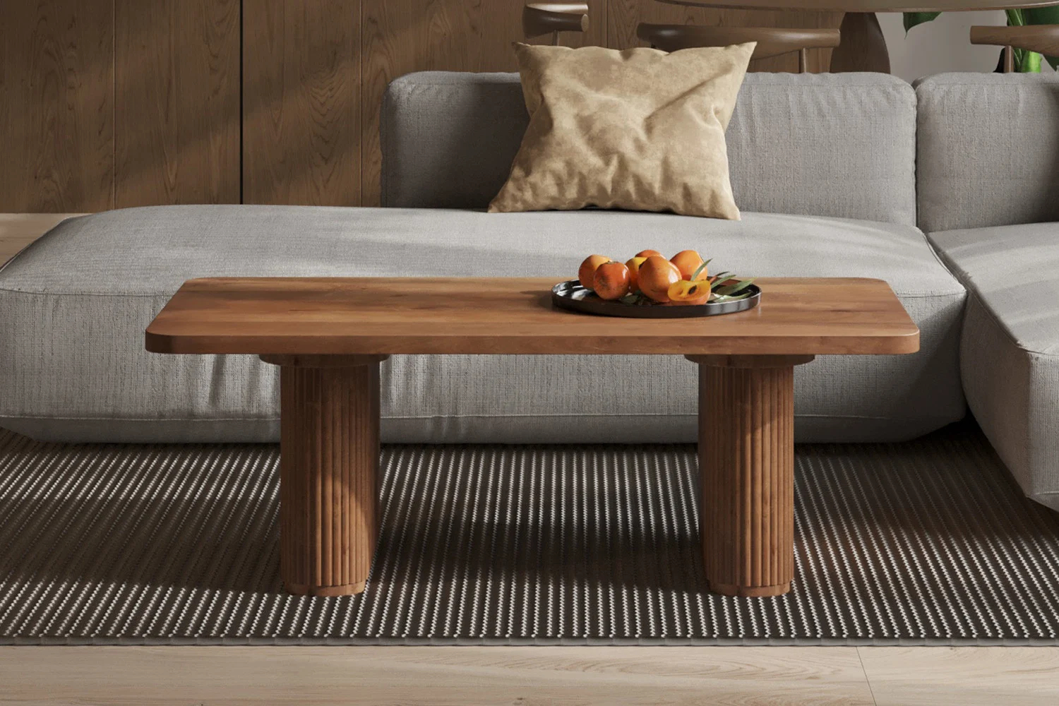 De Salontafel Roest Mangohout Tijdloos, met drie cilindervormige poten, staat op een bruin gestreept vloerkleed voor een lichtgrijze bank met een beige kussen. Een rond dienblad met fruit siert de tafel.