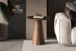 De Salontafel 45 x 50 CM Rond Mangohout, een ronde houten bijzettafel met een taps toelopend onderstel, staat naast een lichtgrijze bank. Hierop liggen een gesloten boek en dennenappels in een moderne, neutraal getinte setting met een groot groen blad aan de linkerkant.