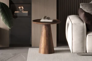 De Salontafel 45 x 50 CM Rond Mangohout, een ronde houten bijzettafel met een taps toelopend onderstel, staat naast een lichtgrijze bank. Hierop liggen een gesloten boek en dennenappels in een moderne, neutraal getinte setting met een groot groen blad aan de linkerkant.