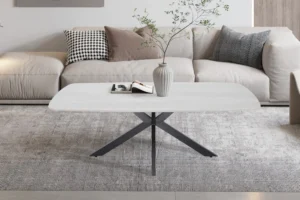 Een moderne woonkamer met een beige bank, gedessineerde en effen kussens, een lichtgrijs vloerkleed en de Salontafel Grijs 130 CM Ovaal Steen als salontafel. Op de tafel staan een gestreepte vaas met takken en een open boek.