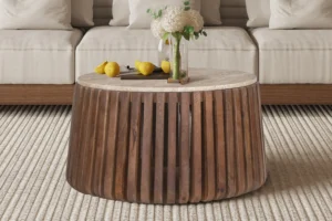 De Salontafel Beige 70 CM Rond Travertin, met een rond travertin design, is vormgegeven met een vaas witte bloemen en drie gele peren, geplaatst op een beige vloerkleed met textuur voor een lichtgekleurde bank.