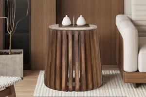 In een moderne woonkamer staat de Bijzettafel 'Luuk'- een 40 cm ronde bijzettafel van mangohout met verticale latten en een blad van beige travertin, versierd met twee kleine witte decoraties. Het staat naast een beige bank op een licht vloerkleed.