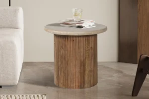 De Bijzettafel 'Louis' 50 cm, gemaakt van mangohout met een beige travertijnen blad, bevat een glas ijswater en een stapel tijdschriften, geplaatst naast een beige bank op een gepolijste vloer.