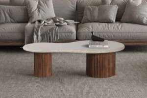 Een moderne woonkamer heeft een grijze bank met een gedrapeerd kleed en de Salontafel Beige 120 CM Travertin, een unieke boonvormige salontafel met een wit blad en twee houten cilinderpoten, waarop twee boeken en een zwarte decoratieve vogel staan.