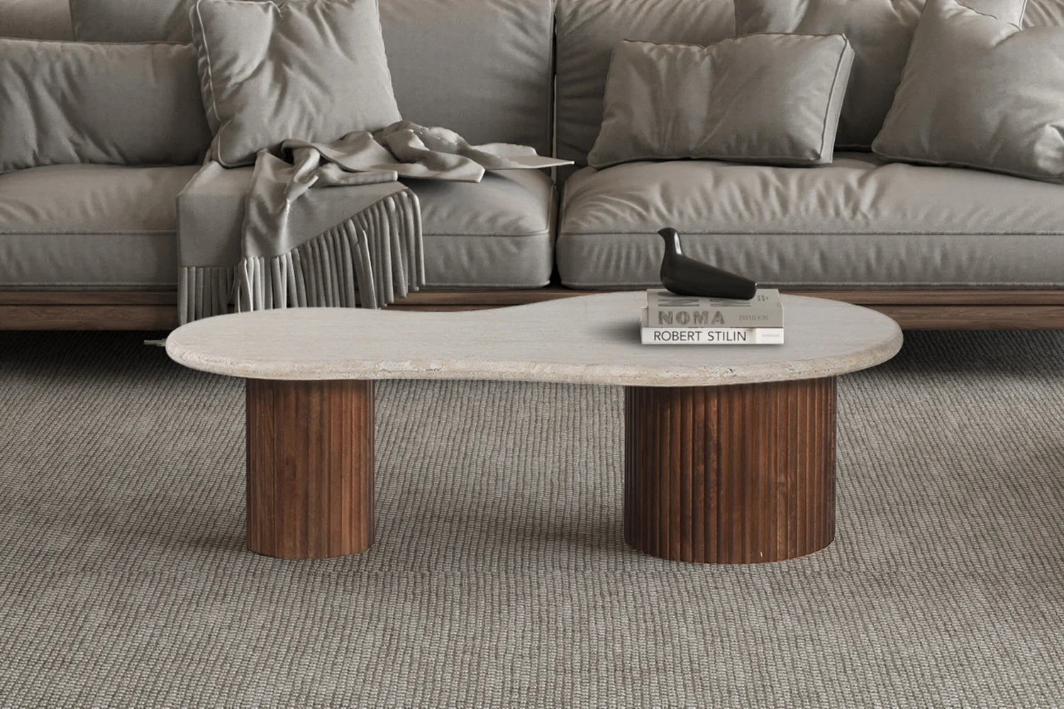 Een moderne woonkamer heeft een grijze bank met een gedrapeerd kleed en de Salontafel Beige 120 CM Travertin, een unieke boonvormige salontafel met een wit blad en twee houten cilinderpoten, waarop twee boeken en een zwarte decoratieve vogel staan.