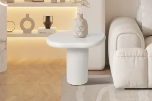 De UITVERKOOP - Salontafel 'Tavira' 58 cm Cement Wit staat naast een crèmekleurige bank op een licht vloerkleed, met daarop een geometrische vaas en gedroogde bloemen, tegen een houten vloer en planken op de achtergrond.
