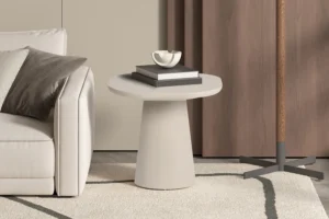 Een moderne woonkamer met de UITVERKOOP - Salontafel 'Tavira' 55 cm Ovaal Cement Beige met twee gestapelde boeken en een kleine schaal, naast een lichte bank en staande lamp op een licht vloerkleed met bruine gordijnen op de achtergrond.
