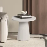 In een moderne woonkamer staat de UITVERKOOP - Salontafel 'Tavira' 55 cm ovaal in cement wit, gestyled met boeken en een decoratieve schaal naast een lichte bank met kussens, een beige vloerkleed, bruine gordijnen en een deel van een staande lamp.