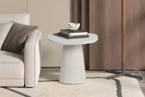 In een moderne woonkamer staat de UITVERKOOP - Salontafel 'Tavira' 55 cm ovaal in cement wit, gestyled met boeken en een decoratieve schaal naast een lichte bank met kussens, een beige vloerkleed, bruine gordijnen en een deel van een staande lamp.