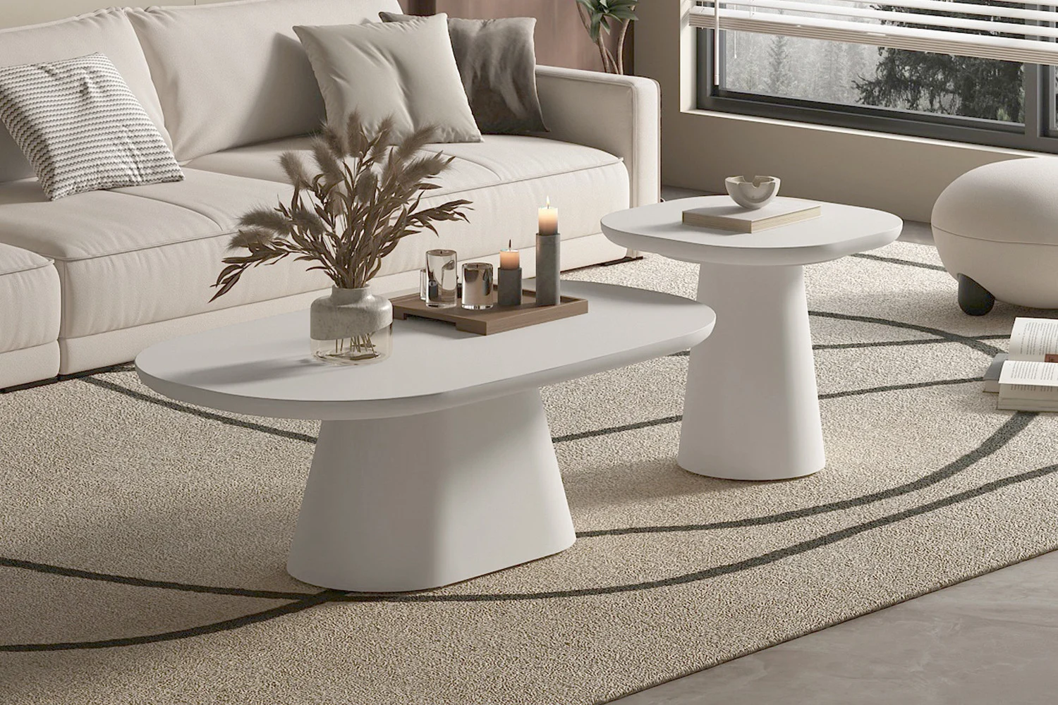 Een moderne woonkamer met een beige bank, een licht vloerkleed en twee ovale 55 cm salontafels van UITVERKOOP - Salontafel Tavira in cementwit. Op de tafels staan kaarsen, een vaas met gedroogde planten en een dienblad bij een groot raam met jaloezieën.