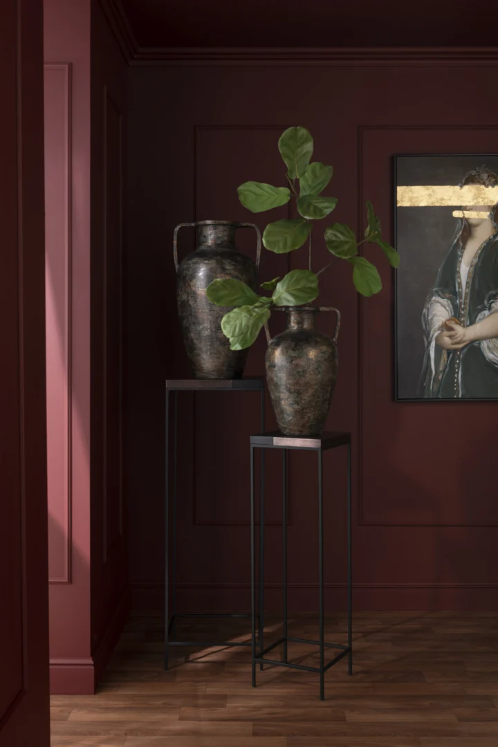 Een decoratieve vaas met groene bladertakken staat op de Bijzettafel Zwart, Brons (41x41 cm, IJzer) naast een donkerrode wand met panelen. Vlakbij hangt een ingelijst portret met een gouden streep boven een houten vloer.