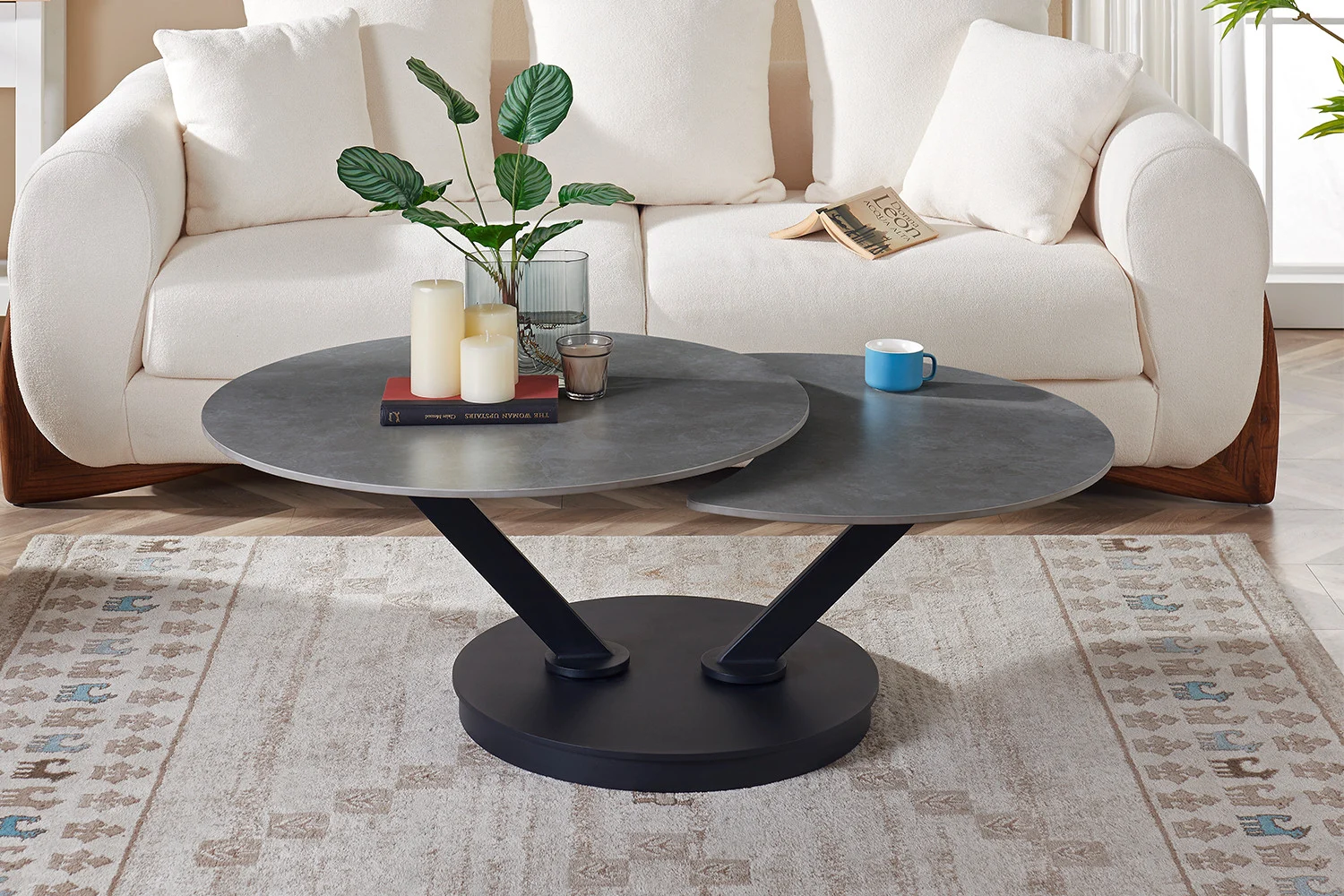 Een moderne woonkamer met een witte bank en de Salontafel Lichtgrijs, Roest Rond Steen met daarop kaarsen, boeken, een plant en een blauwe beker, allemaal op een beige vloerkleed met patroon.