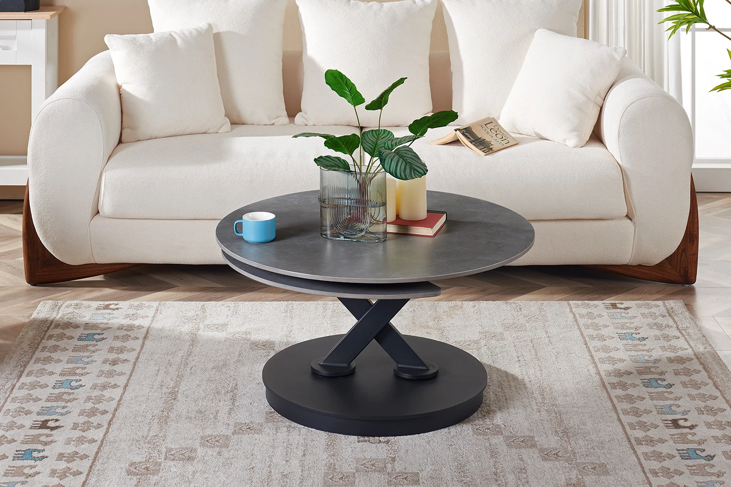 Een moderne woonkamer met een witte bank, de Salontafel Lichtgrijs, Roest Rond Steen salontafel met kruisende zwarte poten, plus een glazen vaas met groene bladeren, een blauw kopje, een boek en een kaars op een vloerkleed met patroon.